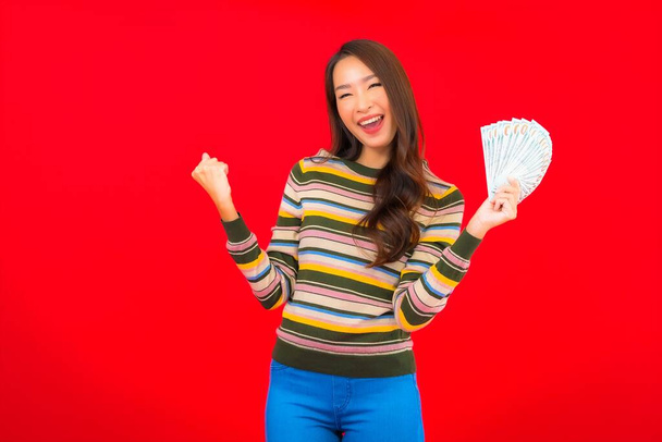 Retrato hermosa joven asiática mujer con dinero en efectivo y teléfono móvil sobre fondo rojo - Foto, Imagen