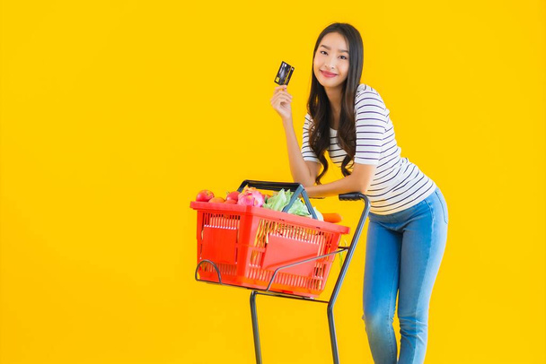 Портрет красива молода азіатська жінка купує продуктовий візок з супермаркету з кредитною карткою на жовтому ізольованому фоні
 - Фото, зображення