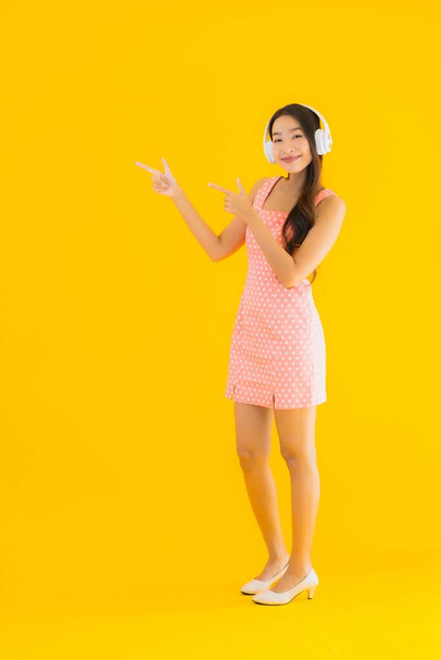 Retrato hermosa joven asiática mujer escuchar música con el teléfono móvil inteligente y auriculares en amarillo aislado fondo
 - Foto, Imagen