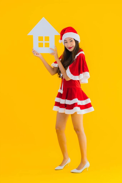 Porträt schöne junge asiatische Frau tragen Weihnachtskleidung und Hut zeigen nach Hause Hausschild mit einer Menge Bargeld und Geld auf gelbem Hintergrund isoliert - Foto, Bild