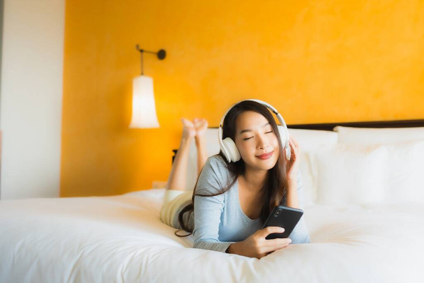 Πορτρέτο όμορφη νεαρή Ασιάτισσα γυναίκα χρησιμοποιώντας κινητό τηλέφωνο με ακουστικά για να ακούσετε μουσική στο εσωτερικό του υπνοδωματίου - Φωτογραφία, εικόνα