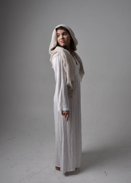 Ganztägiges Porträt einer jungen Frau in klassischem weißen Kleid und Kopftuch im biblischen Stil, stehend auf hellem Studiohintergrund. - Foto, Bild