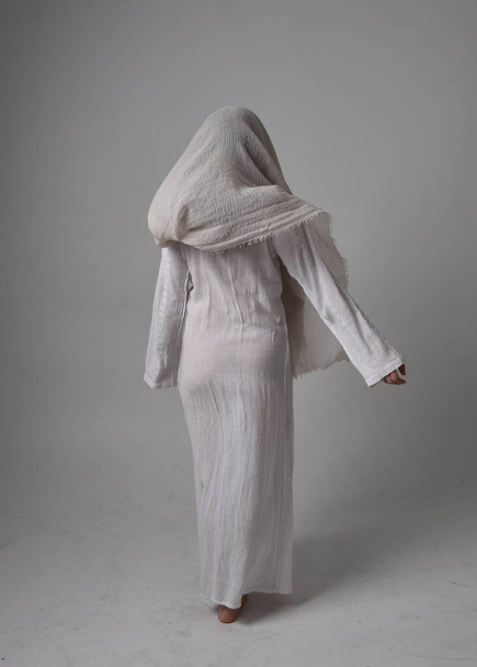 Retrato de comprimento total da jovem mulher vestindo vestido branco clássico e um véu de cobertura da cabeça em estilo bíblico, em pé posar no fundo do estúdio de luz. - Foto, Imagem