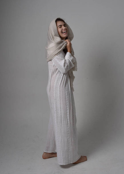 Ganztägiges Porträt einer jungen Frau in klassischem weißen Kleid und Kopftuch im biblischen Stil, stehend auf hellem Studiohintergrund. - Foto, Bild
