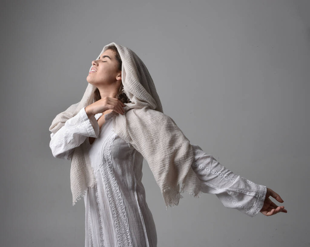 close up  portrait of young woman wearing classical white gown and a head covering veil in biblical style, standing worshiping  pose on light studio background. - Фото, изображение