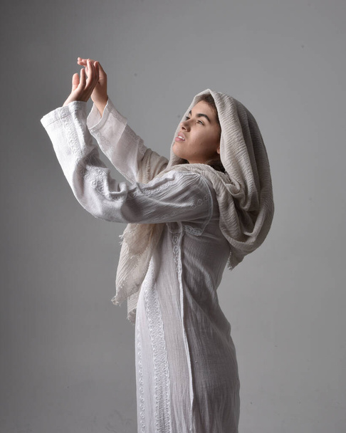 portrait en gros plan d'une jeune femme portant une robe blanche classique et un voile couvrant la tête dans un style biblique, posant debout vénérant sur fond de studio léger. - Photo, image