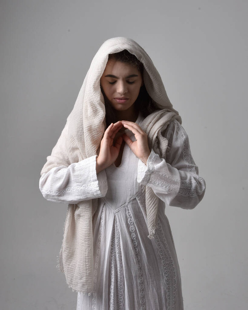 close up retrato de jovem mulher vestindo vestido branco clássico e uma cabeça cobrindo véu em estilo bíblico, em pé adorando pose no fundo do estúdio de luz. - Foto, Imagem