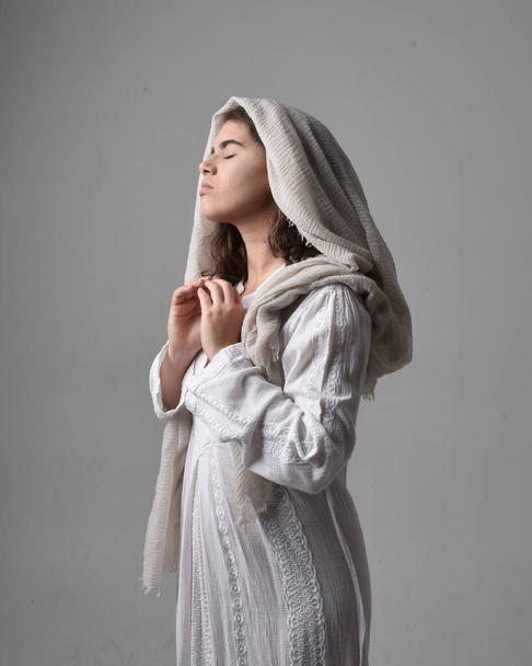 close up  portrait of young woman wearing classical white gown and a head covering veil in biblical style, standing worshiping  pose on light studio background. - Photo, Image