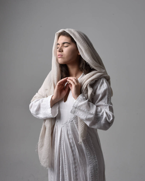 close up retrato de jovem mulher vestindo vestido branco clássico e uma cabeça cobrindo véu em estilo bíblico, em pé adorando pose no fundo do estúdio de luz. - Foto, Imagem