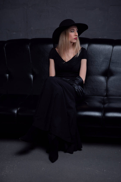 sexy long-haired blonde with bright makeup in a black hat and a stylish elegant evening dress sits on a black leather sofa - Фото, изображение
