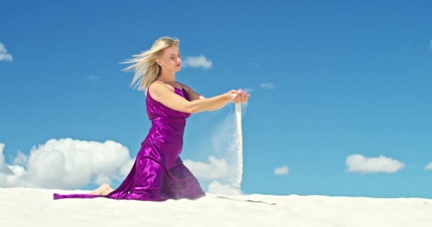 Cinemática cámara lenta de una mujer que sacó arena de sus manos mientras estaba sentada en el desierto. Mujer descalza viajera en vestido balanceándose en el viento sobre la superficie montañosa del desierto de arena con una nube en - Imágenes, Vídeo