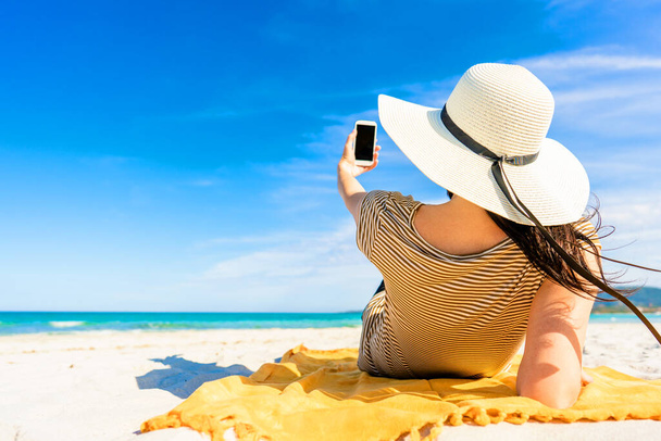 Обратный снимок модной молодой неузнаваемой женщины в большой белой шляпе, лежащей на песке тропического пляжа и делающей селфи со смартфоном, делящимся отпуском в социальной сети. Яркая цветная фотография - Фото, изображение