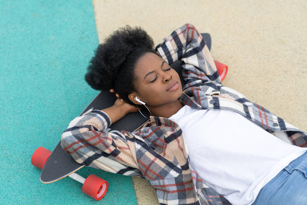 Chica africana relajada acostada en longboard escuchar música en auriculares inalámbricos con los ojos cerrados al aire libre - Foto, imagen
