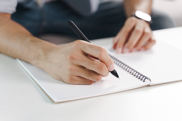 Крупный план молодого человека в повседневной ткани руки, записанные на блокноте, ноутбук с шариковой ручкой на столе. - Фото, изображение