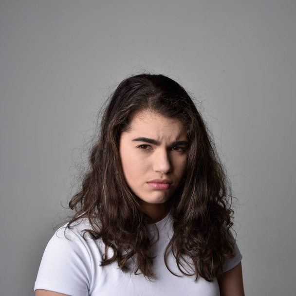 Κοντινό πορτραίτο νεαρής γυναίκας με φυσικά καστανά μαλλιά, πάνω από τις κορυφαίες εκφράσεις του προσώπου σε ανοιχτό γκρι φόντο στούντιο. - Φωτογραφία, εικόνα