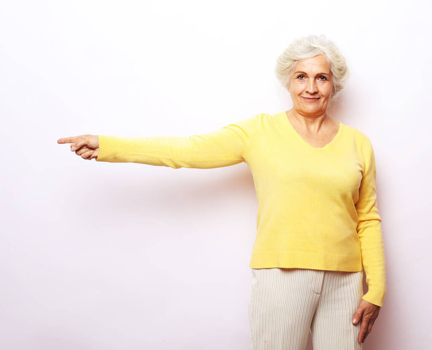 Portret van oude vrouw in gele trui demonstreren, kijken, wijzen met de vingers omhoog om ruimte te kopiëren. - Foto, afbeelding