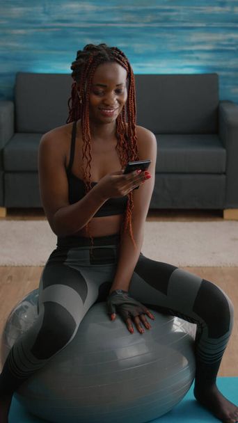 Slim fit μαύρη γυναίκα κάθεται στο fitness ball πληκτρολογώντας μήνυμα στο τηλέφωνο - Φωτογραφία, εικόνα