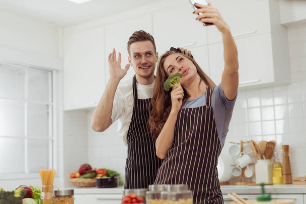 Genç çift mutfakta birlikte yemek yaparken selfie çekmeyi sever. Mutfak odasındaki arkadaşlarıyla video görüşmesi yapan mutlu bir adam ve kadın.. - Fotoğraf, Görsel