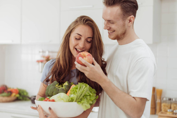 Young man giving apple to his girlfriend while cooking together in the kitchen. Happy couple love cooking together at home. - Foto, afbeelding
