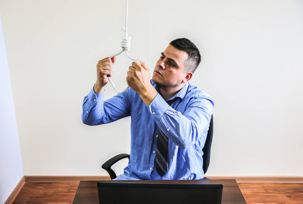 Noose is above the office worker. Suicide concept. Hanging because of work stress. Depression of burnout. Terrible life situation. Man near the laptop at the desk. - 写真・画像