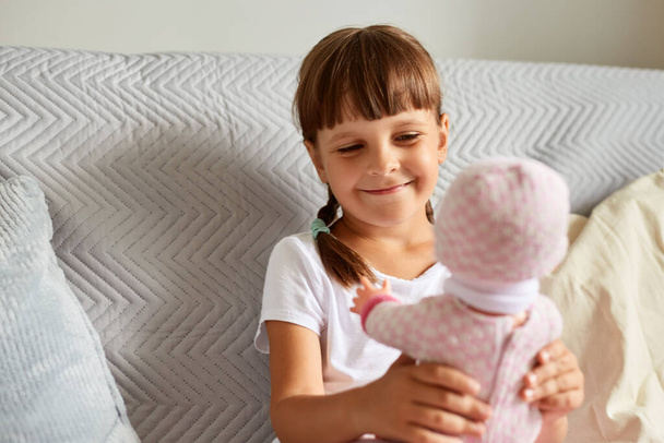 Smiling happy charming kid with dark hair and pigtails holding in her hand a doll, looking at toy with smile, kid wearing white casual t shirt playing indoor. - 写真・画像