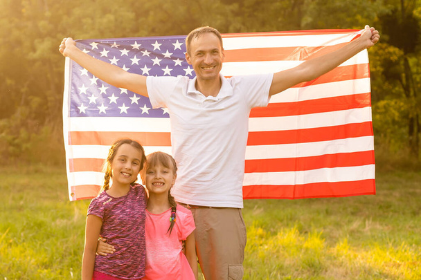 幸せな家族が裏庭で彼らの後ろにアメリカの旗を持って一緒に座っている。アメリカ独立記念日を祝う子供たちと笑顔のカップルアメリカ国旗を持って - 写真・画像