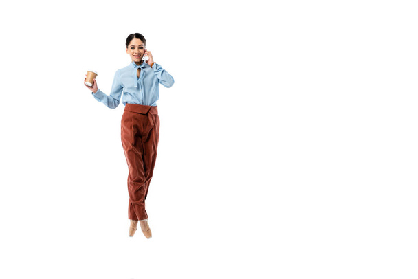 Uśmiechnięta balerina rozmawiająca przez telefon komórkowy i trzymająca papierowy kubek na białym  - Zdjęcie, obraz
