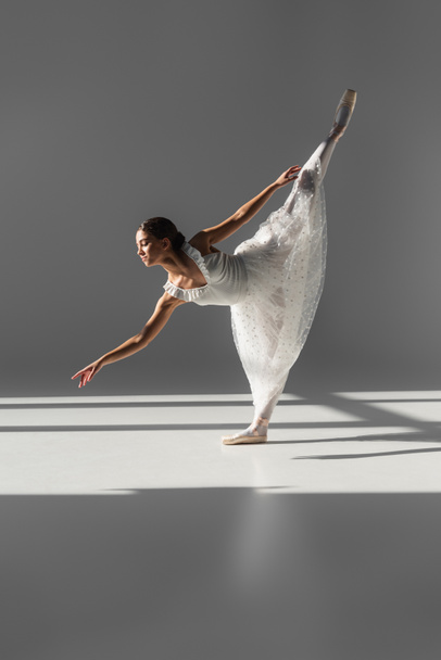 Side view of ballerina standing on one leg on grey background  - Zdjęcie, obraz