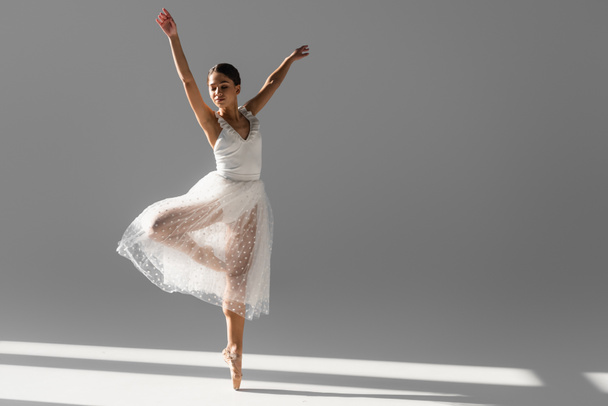 Brunette ballerina in white skirt dancing with closed eyes on grey background with sunlight - Photo, Image