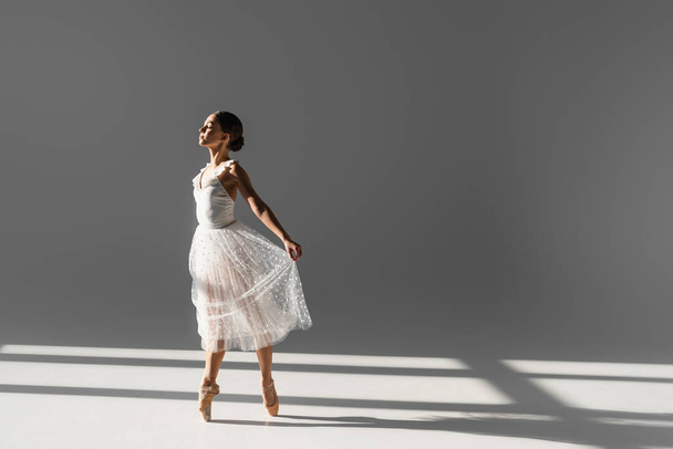 Πλευρική άποψη της μπαλαρίνας κρατώντας φούστα, ενώ χορεύουν σε γκρι φόντο με το φως του ήλιου - Φωτογραφία, εικόνα