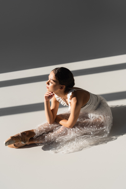 Vista lateral da bailarina flexão enquanto sentado sobre fundo cinza com luz solar - Foto, Imagem