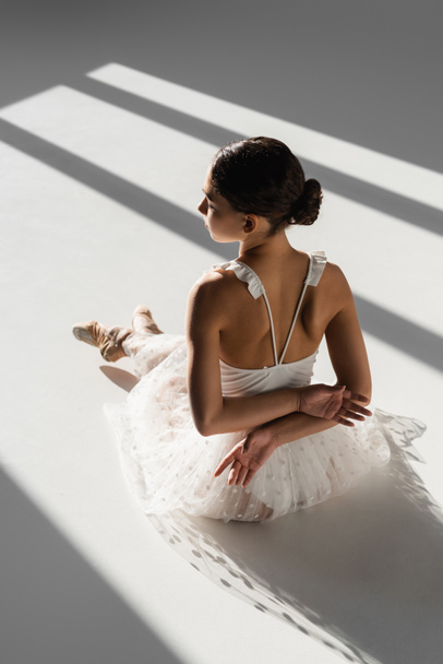 Side view of ballerina in white dress and pointe shoes sitting in sunlight on grey background  - Fotoğraf, Görsel