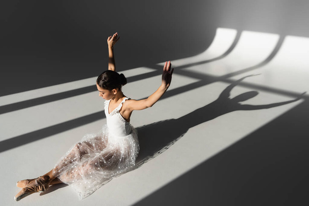 Boční pohled na balerína zvedání rukou při sezení na šedém pozadí se slunečním světlem - Fotografie, Obrázek