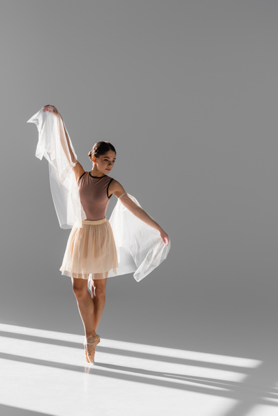 Hübsche Ballerina in beigem Rock, die mit Tuch auf grauem Hintergrund mit Sonnenlicht tanzt - Foto, Bild