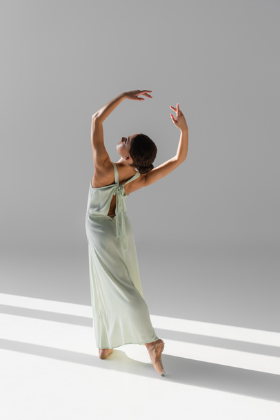 Side view of young dancer in pointe shoes standing on grey background with sunlight - Foto, imagen