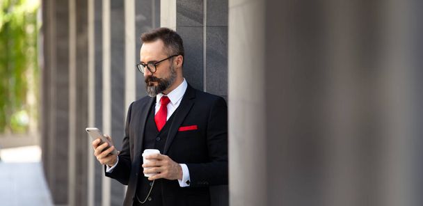 Handsome Caucasian business man using mobile phone and holding take away coffee cup outside apartment in the morning before going to work with copy space - Photo, Image