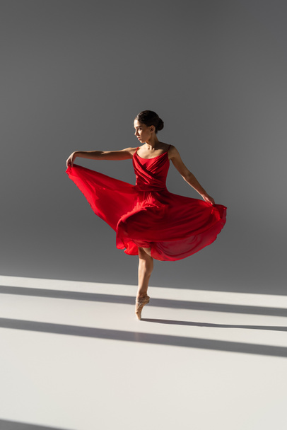 Vista lateral de bailarina bailando y sosteniendo vestido rojo sobre fondo gris con luz solar - Foto, Imagen
