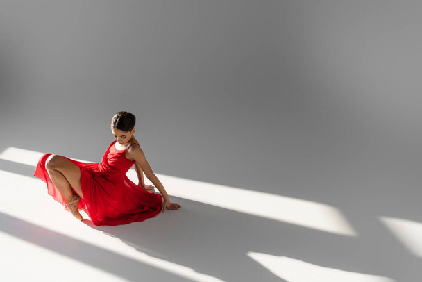 Ballerina in red dress dancing on floor on grey background with sunlight - Foto, Imagen