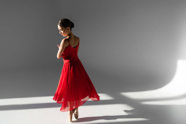 Боковой вид профессиональной балерины танцующей в красном платье на сером фоне с солнечным светом - Фото, изображение