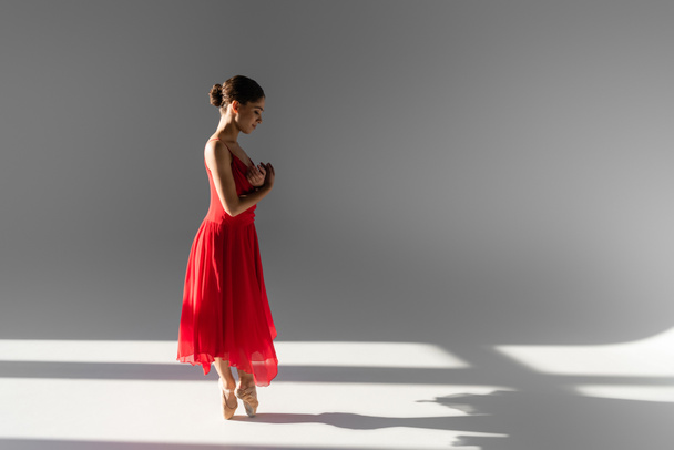 Боковой вид молодой балерины, танцующей в красном платье и пуантах на сером фоне с солнечным светом - Фото, изображение