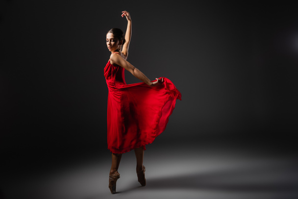 Pretty ballerina holding red dress while dancing on black background  - Фото, изображение