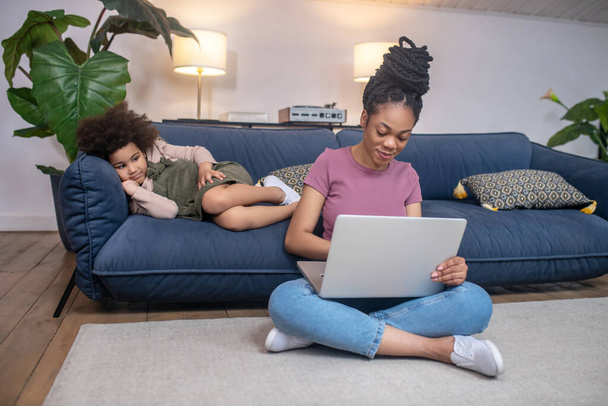 Woman with laptop on floor and daughter on couch - Photo, image