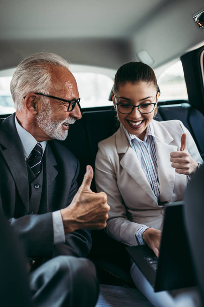 Good looking senior business man and his young woman colleague or coworker sitting on backseat in luxury car. They talking, smiling and using laptop and smart phones. Transportation in corporate business concept. - Фото, изображение