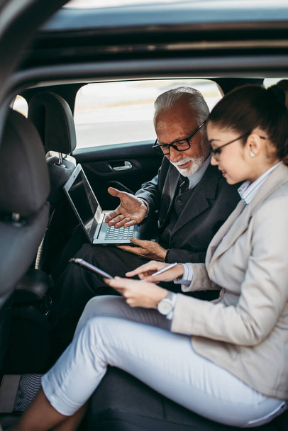 Good looking senior business man and his young woman colleague or coworker sitting on backseat in luxury car. They talking, smiling and using laptop and smart phones. Transportation in corporate business concept. - Fotoğraf, Görsel