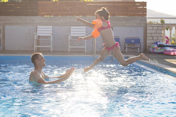 father catches little girl jumping in pool against sea - Photo, Image