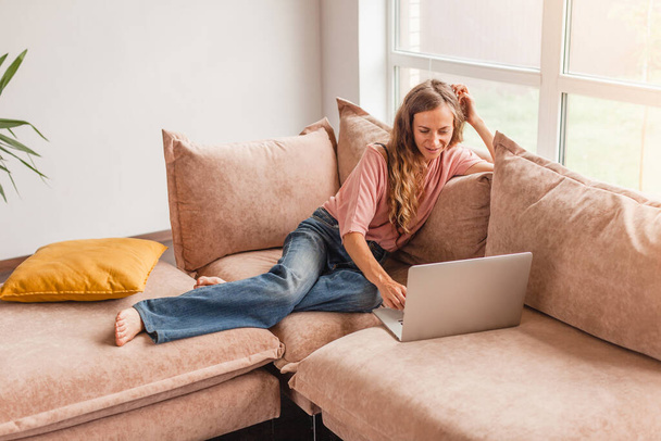 Beautiful cheerful woman using silver laptop while sitting on sofa in living room at home - Φωτογραφία, εικόνα