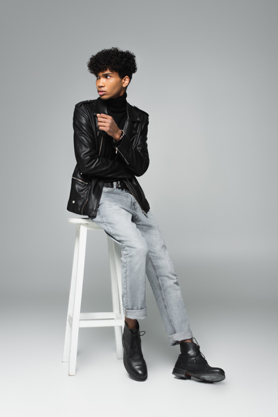 young african american man in leather jacket and jeans looking away on high stool on grey background - Foto, afbeelding