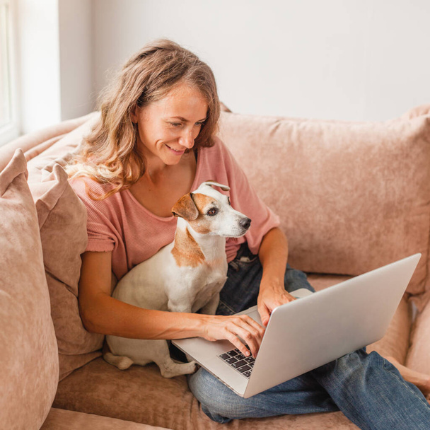 Young woman is using laptop pc for remote conversation with friends. Funny girl with dog having fun staying at home - Fotografie, Obrázek