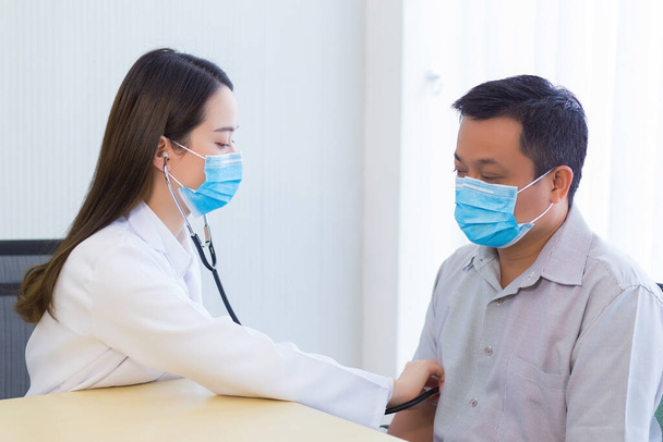 Aasialainen miespotilas tarkistetaan hänen terveytensä, kun taas naislääkäri käyttää stetoskooppia kuullakseen sykkeen ja hänen äänensä Coronavirus-pandemiassa pitämällä kirurgista naamiota kaikkina aikoina.. - Valokuva, kuva