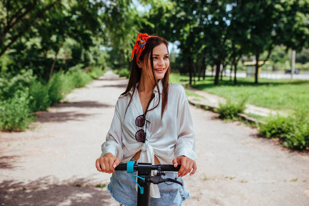かわいいブルネットの女の子は彼女が白いシャツを着ている公園の電極スクーターに乗っています。エコ輸送と家賃の概念は.  - 写真・画像